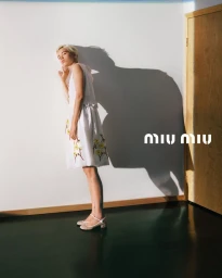 Model with blonde hair wearing a white floral dress standing against a plain wall casting a large shadow