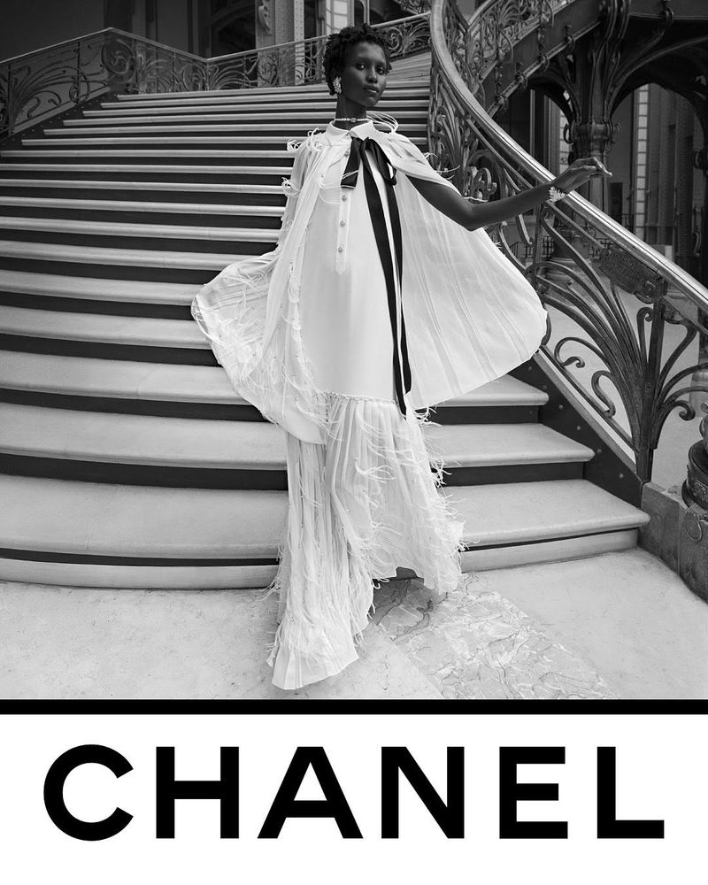 Elegant fashion shot of a woman in a white gown posing on a grand staircase with Chanel logo at the bottom. Black and white aesthetic.