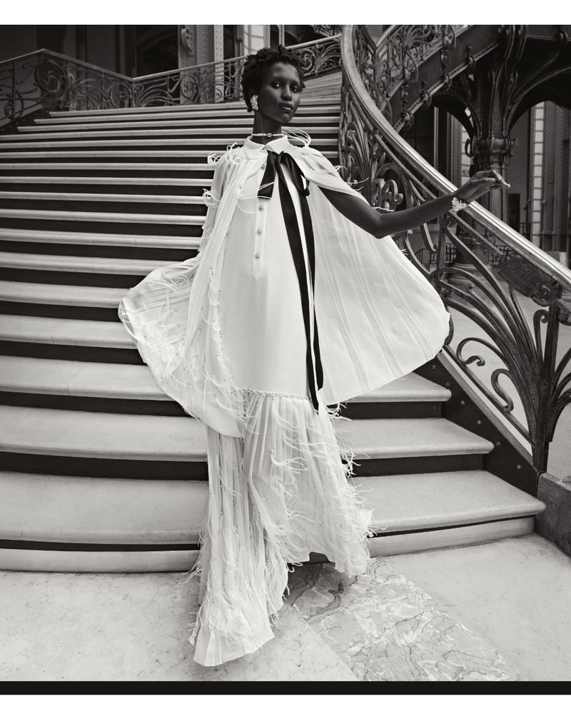 Elegant black and white fashion shot of an adult woman in a stylish cape dress descending a grand staircase in a luxurious interior.