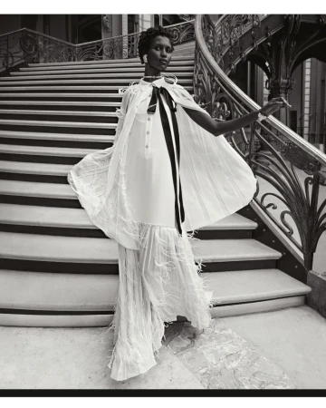 Elegant black and white fashion shot of an adult woman in a stylish cape dress descending a grand staircase in a luxurious interior.