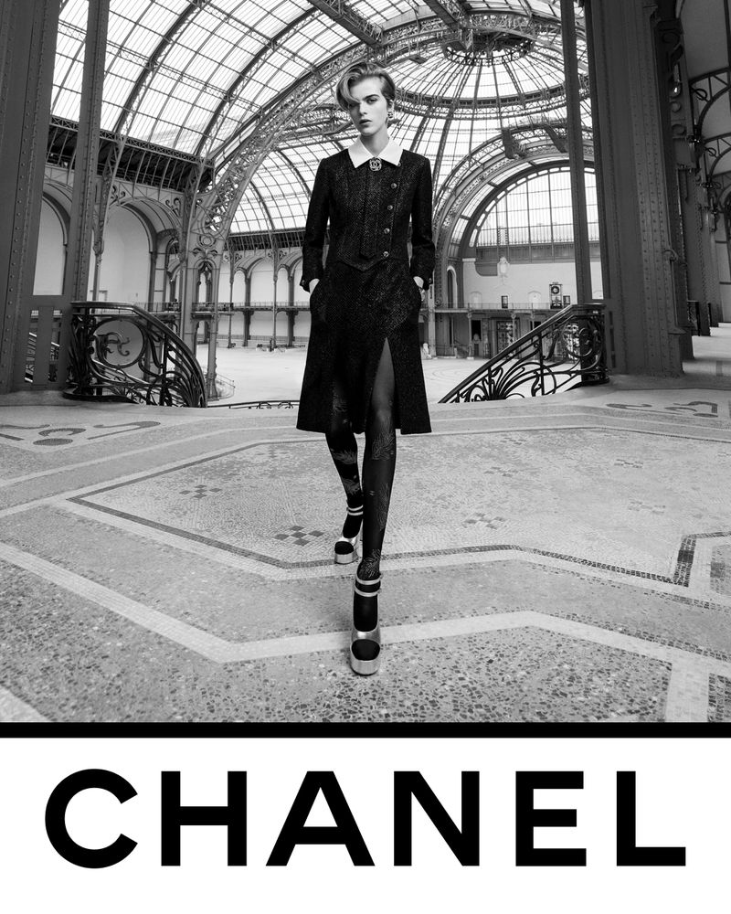 Fashionable woman in black coat, stockings, and platform shoes strides through an ornate building atrium, striking a confident pose for Chanel.