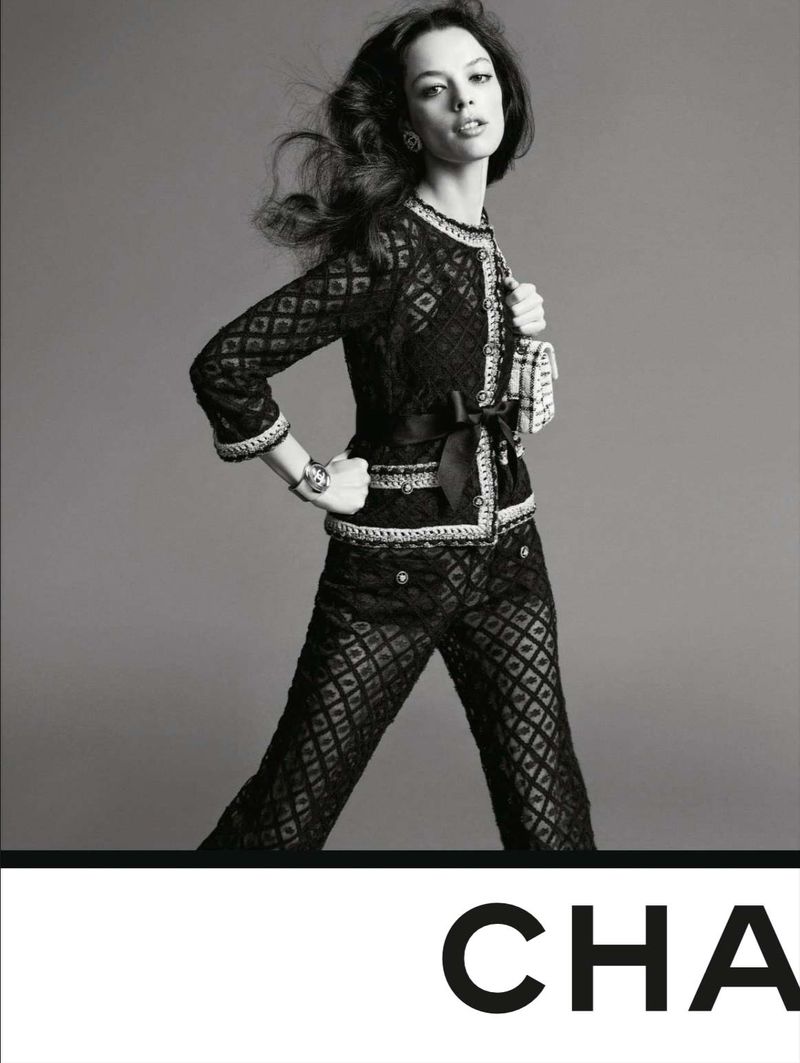 An adult woman in a dark patterned suit is posing in a studio, showcasing a luxury fashion brand and wearing dark makeup.