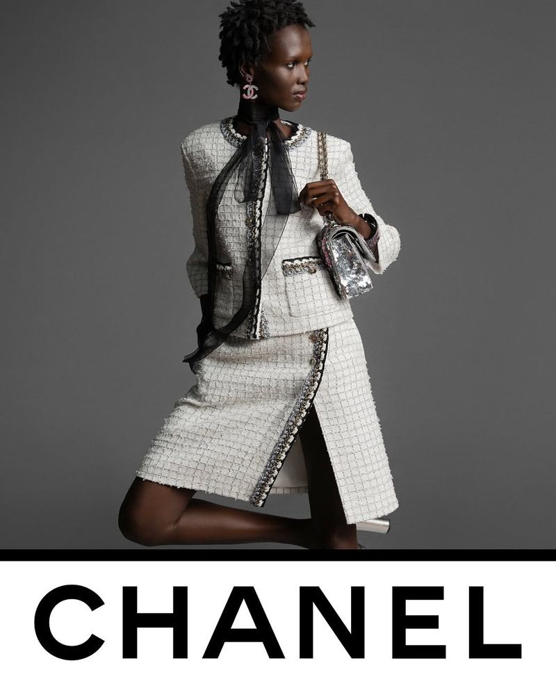 Elegant Black woman posing in white tweed skirt suit with silver purse, wearing Chanel logo earrings, against a neutral grey backdrop.