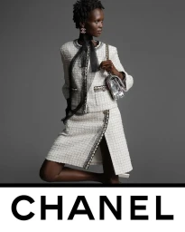 Model wearing a white tweed suit with a black bow and holding a metallic handbag against a solid grey background