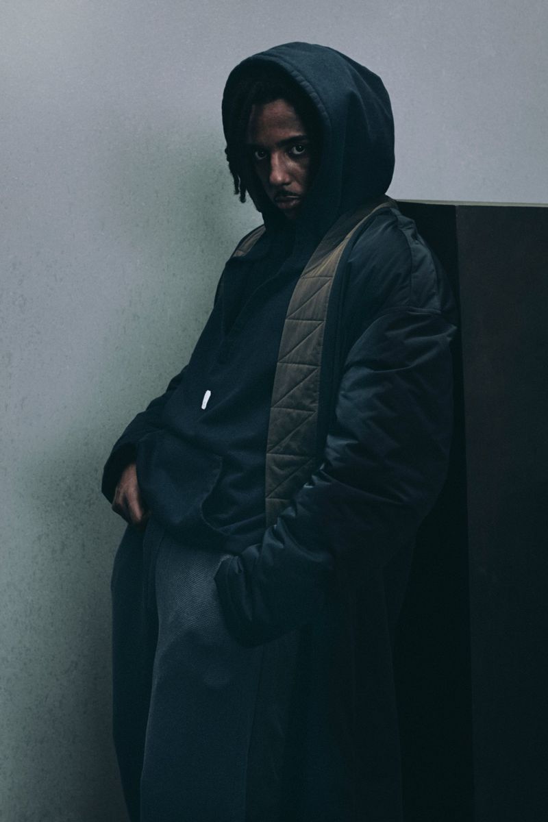 Man wearing a dark hooded jacket and layered coat posing against a plain grey studio backdrop