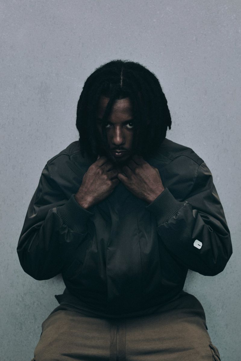 Portrait of adult male in a dark green bomber jacket and tan pants sitting against a textured gray wall. Dark hair.