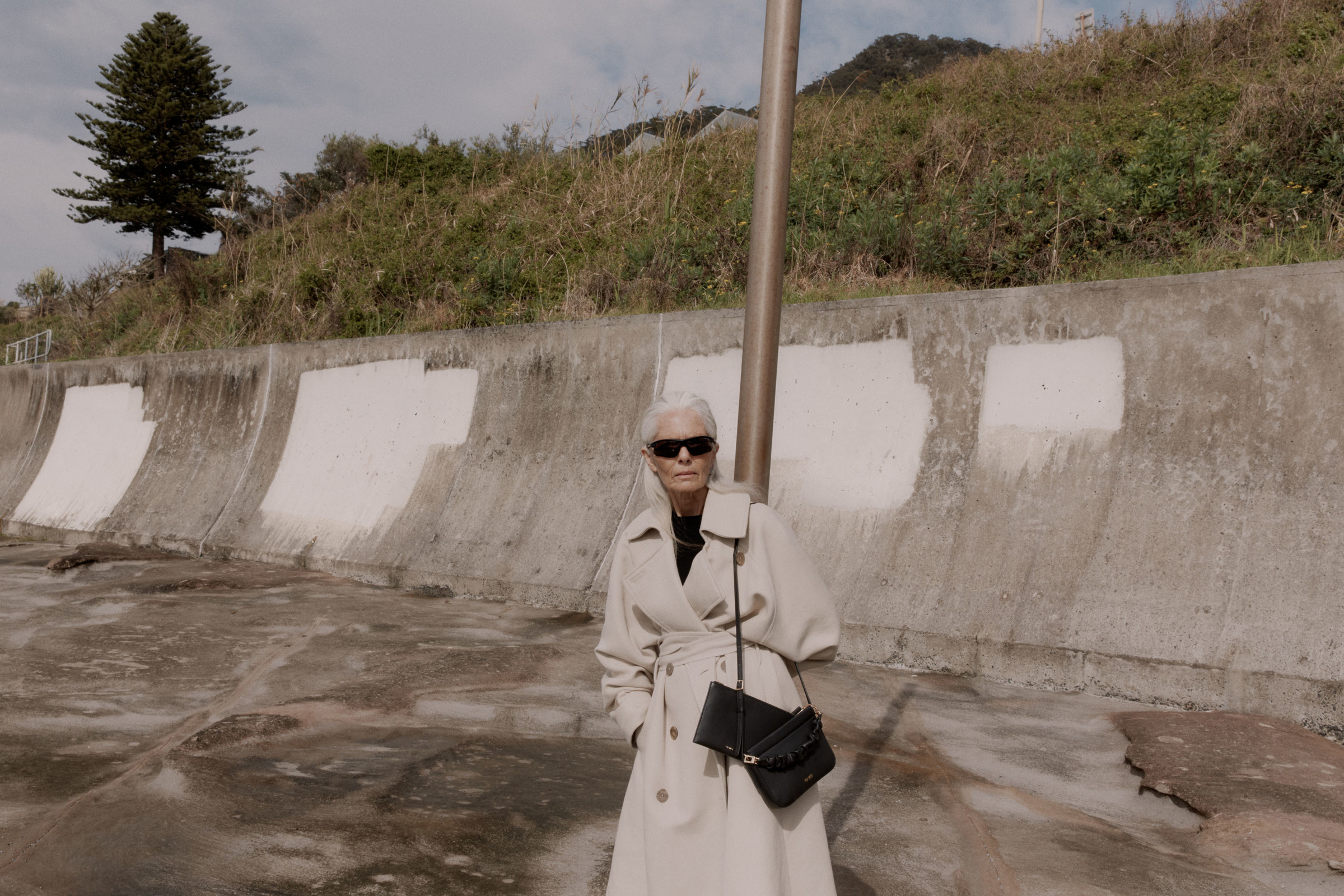Elderly woman poses in a beige coat and sunglasses with a black purse against a concrete wall on a cloudy day outdoors.