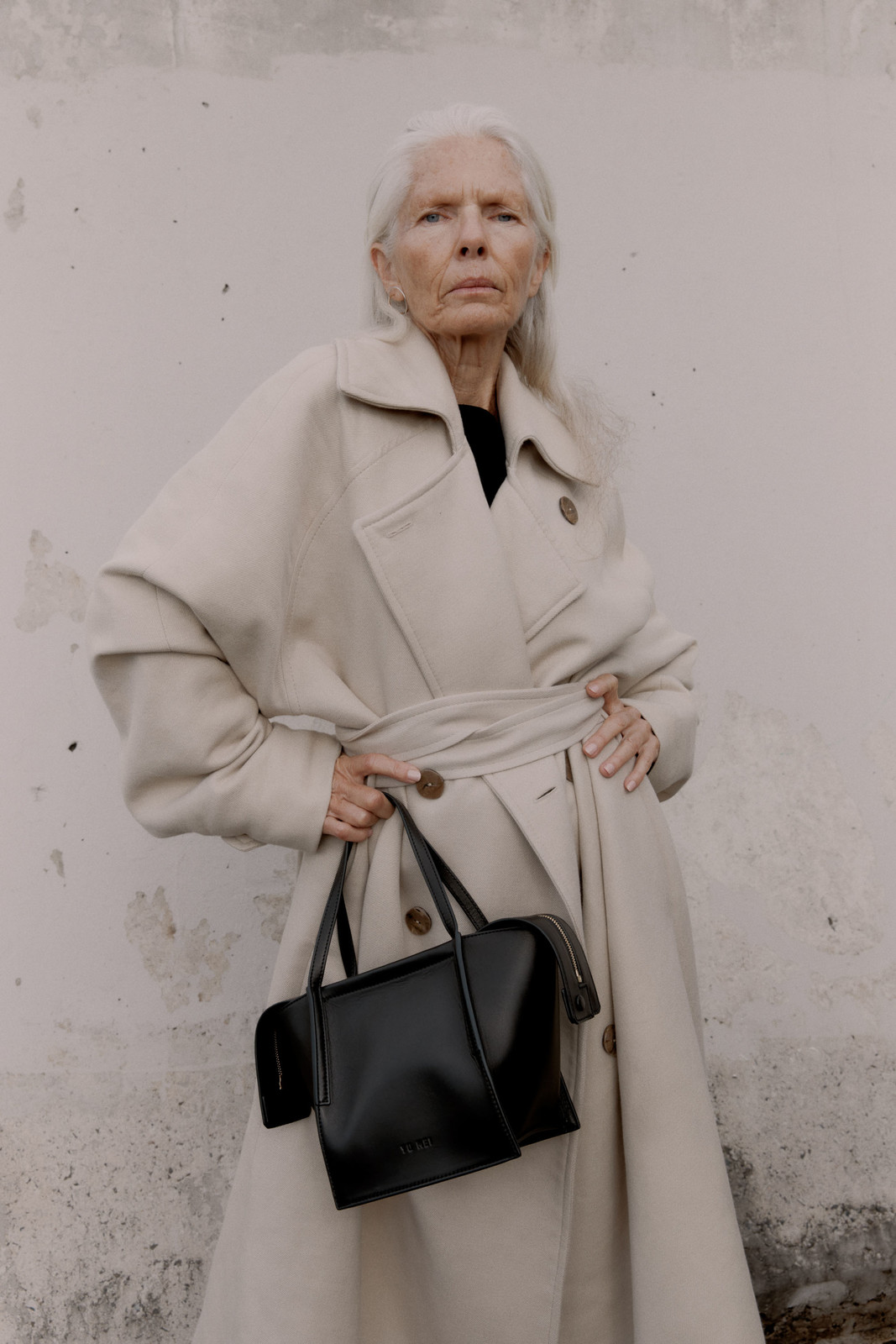 Elderly woman in a beige trench coat poses with a black leather handbag against a light wall, fashion photography.