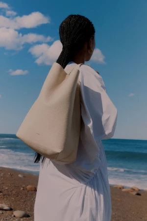 Woman on beach carrying a large beige tote bag. She is wearing a white dress. The background features the ocean and cloudy sky.