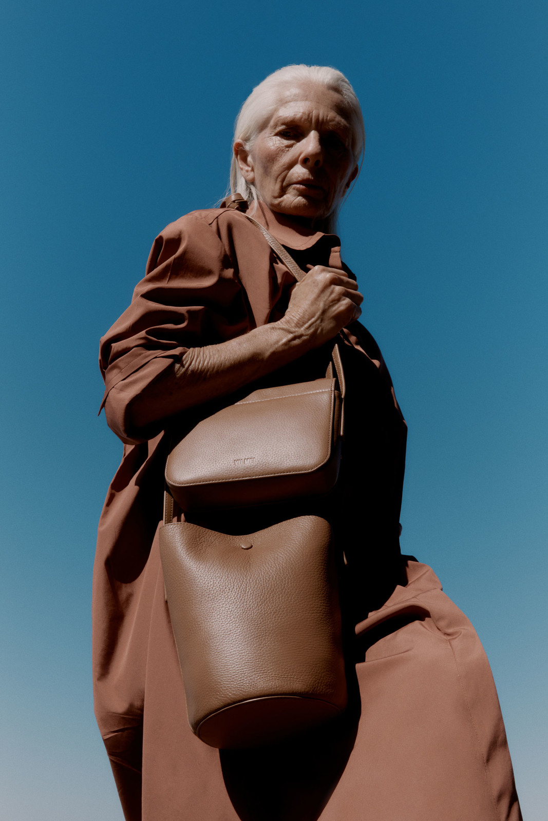 Elderly woman in brown coat with a brown purse and blue sky background, low angle. Fashion, accessory, and clothing.