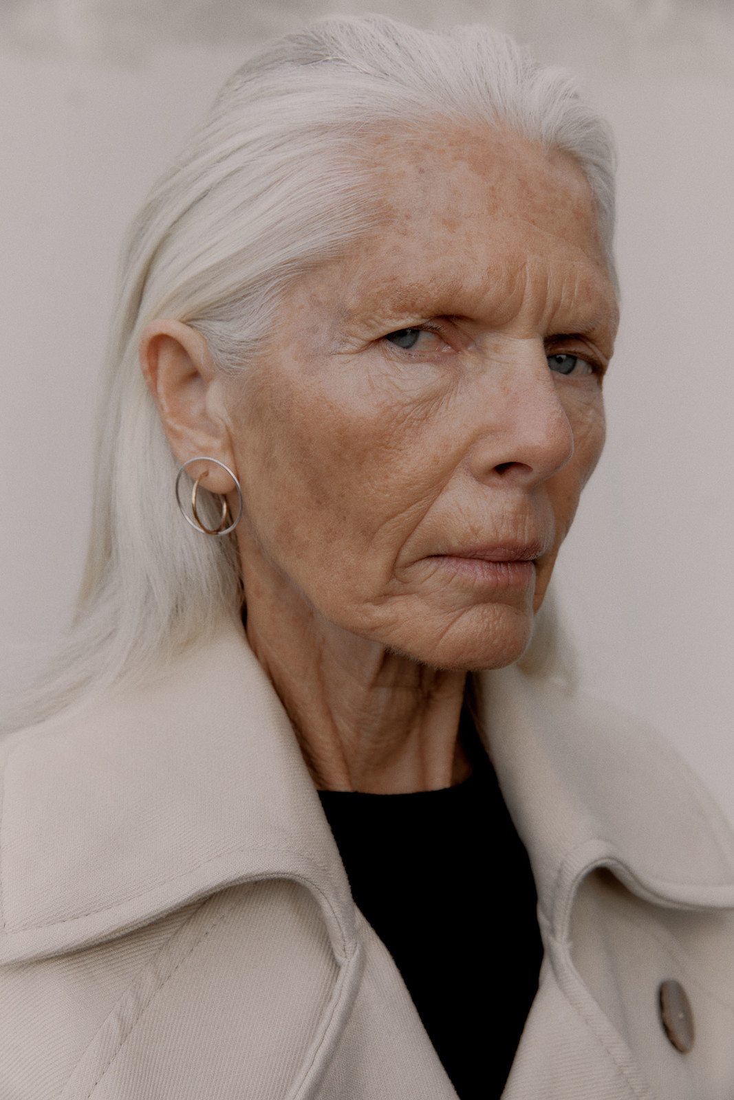 Close-up portrait of an elderly woman with long, white hair, wearing a tan jacket and hoop earrings, set against a neutral background.