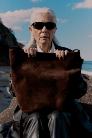 Elderly woman with sunglasses and a brown tote bag posing outdoors on a beach with rocks, sea, and sky visible in the background.
