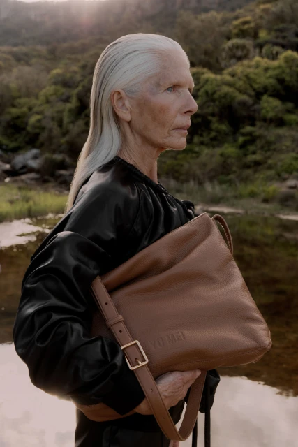 Elegant mature woman with grey hair in a black outfit holds a brown leather handbag in a natural outdoor setting by water.