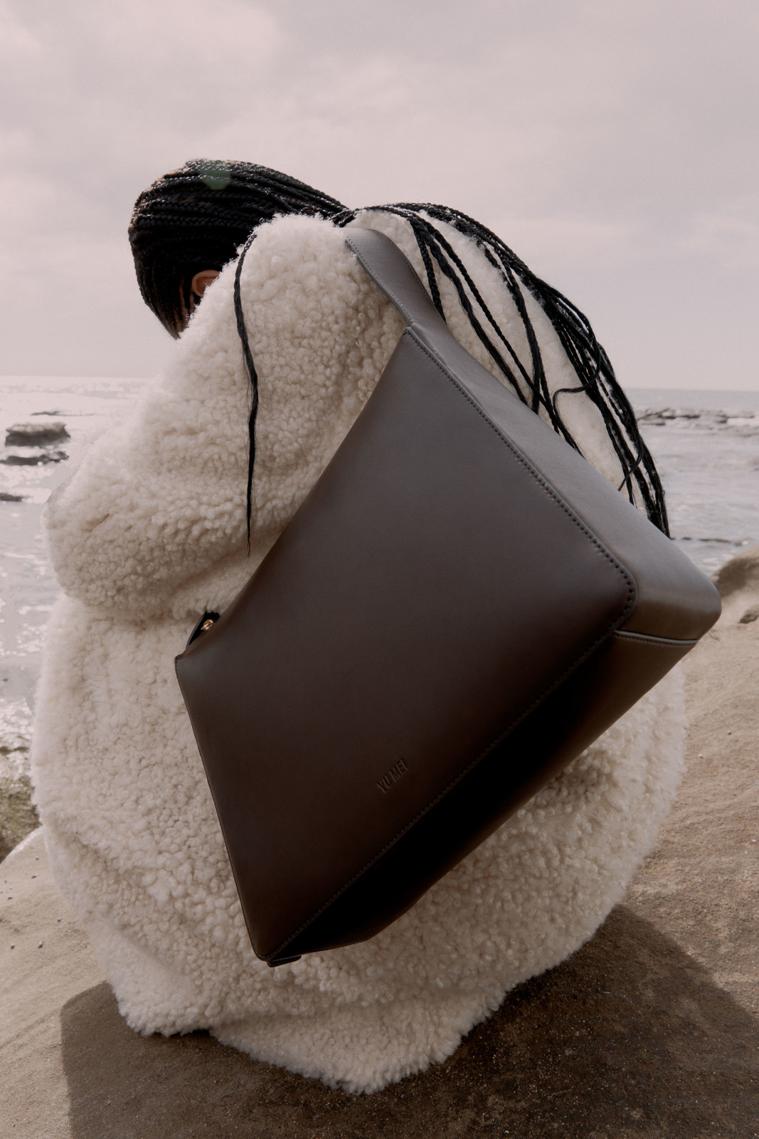 Fashion shot of a person with braids wearing a sheepskin coat and carrying a brown leather tote bag near the ocean shore.