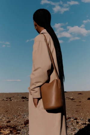 Model with long braided hair wearing a beige coat and shoulder bag outdoors against a blue sky