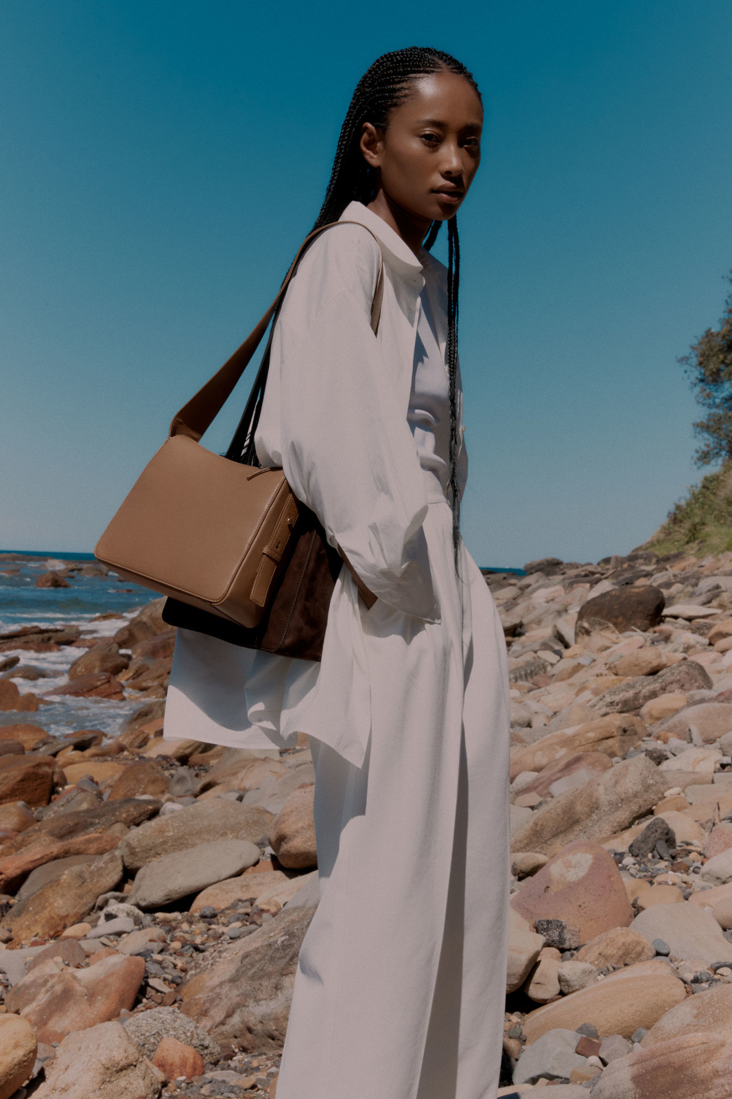 A stylish woman poses on a rocky beach with a leather messenger bag against a backdrop of blue sky and ocean.
