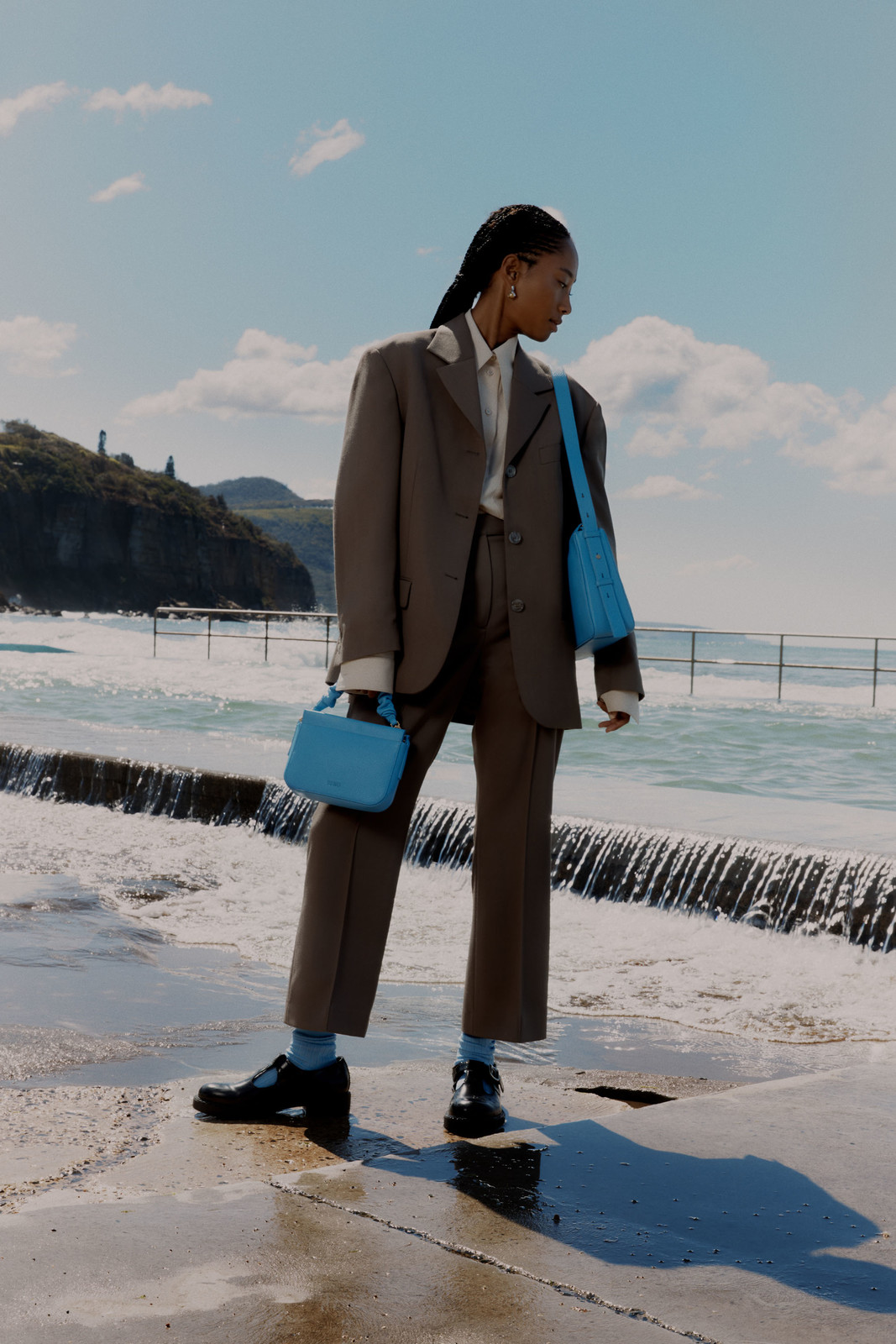 Model in a brown suit holding two light blue handbags standing near an ocean pool under a clear sky.