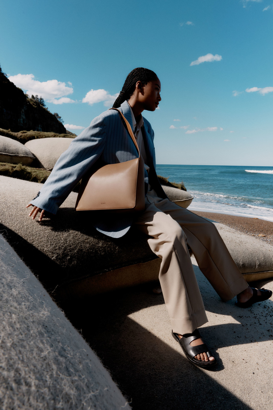 Fashion shoot of a Black woman in a blue jacket and tan pants, wearing a tan leather purse, seated outdoors with the ocean in the background.