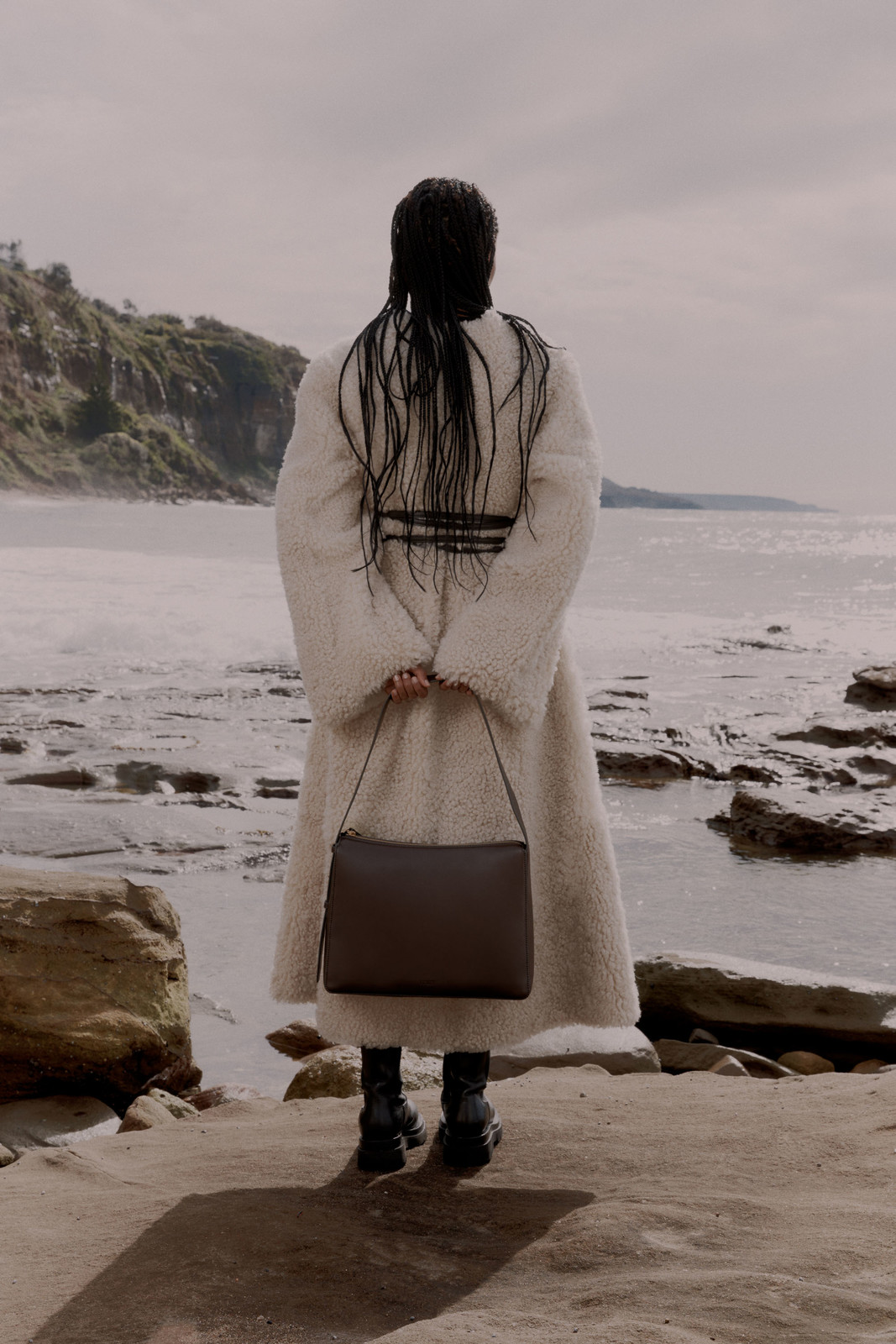 Model standing on a rocky coast wearing a cream coat and holding a dark handbag