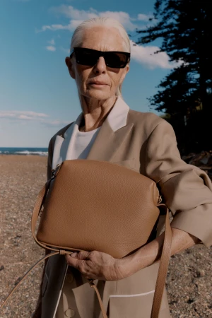 Older woman with white hair wearing sunglasses and a tan blazer holding a brown leather handbag outdoors by the coast