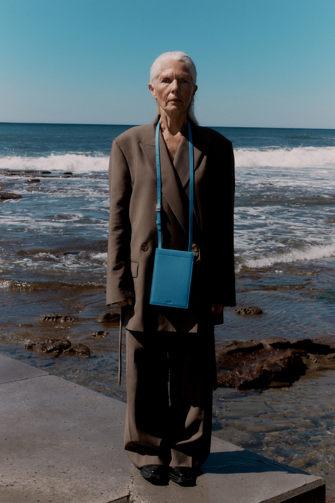 Older woman with silver hair wearing a loose brown pantsuit and a bright blue crossbody bag near the ocean