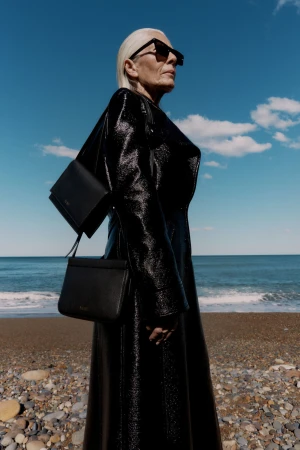 Older woman with silver hair wearing a black patent leather coat and sunglasses on a pebble beach with ocean background
