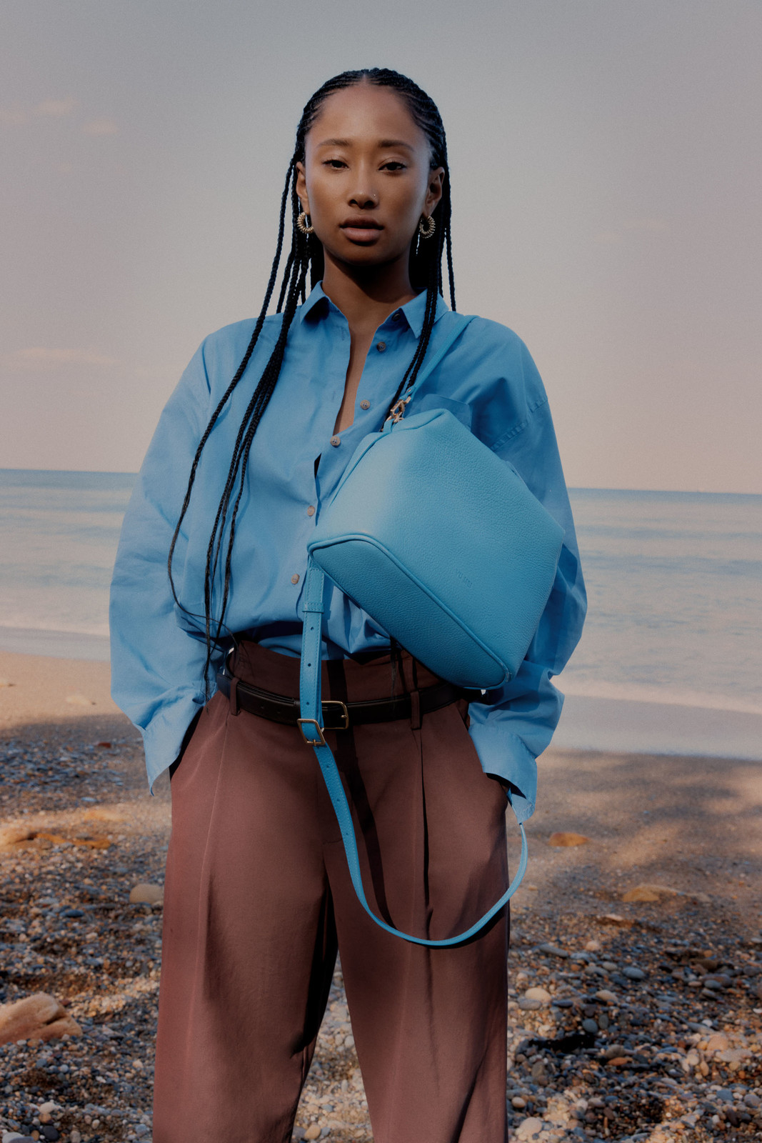 A young Asian woman poses on a beach with a light blue bag, wearing a light blue shirt and brown pants against a seascape backdrop.