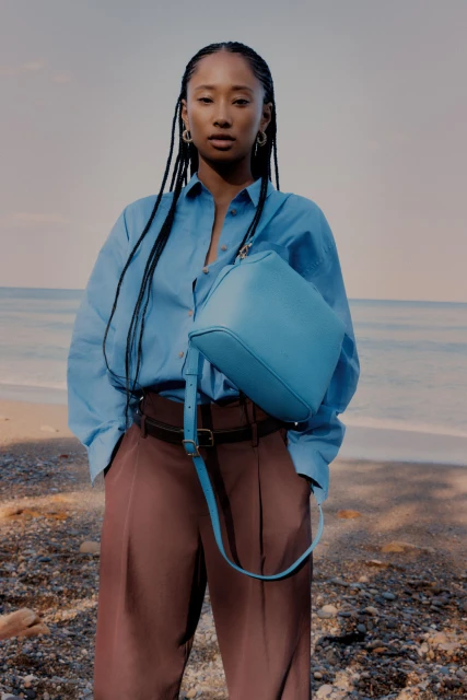 A young Asian woman poses on a beach with a light blue bag, wearing a light blue shirt and brown pants against a seascape backdrop.