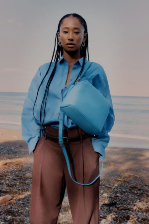 A young Asian woman poses on a beach with a light blue bag, wearing a light blue shirt and brown pants against a seascape backdrop.