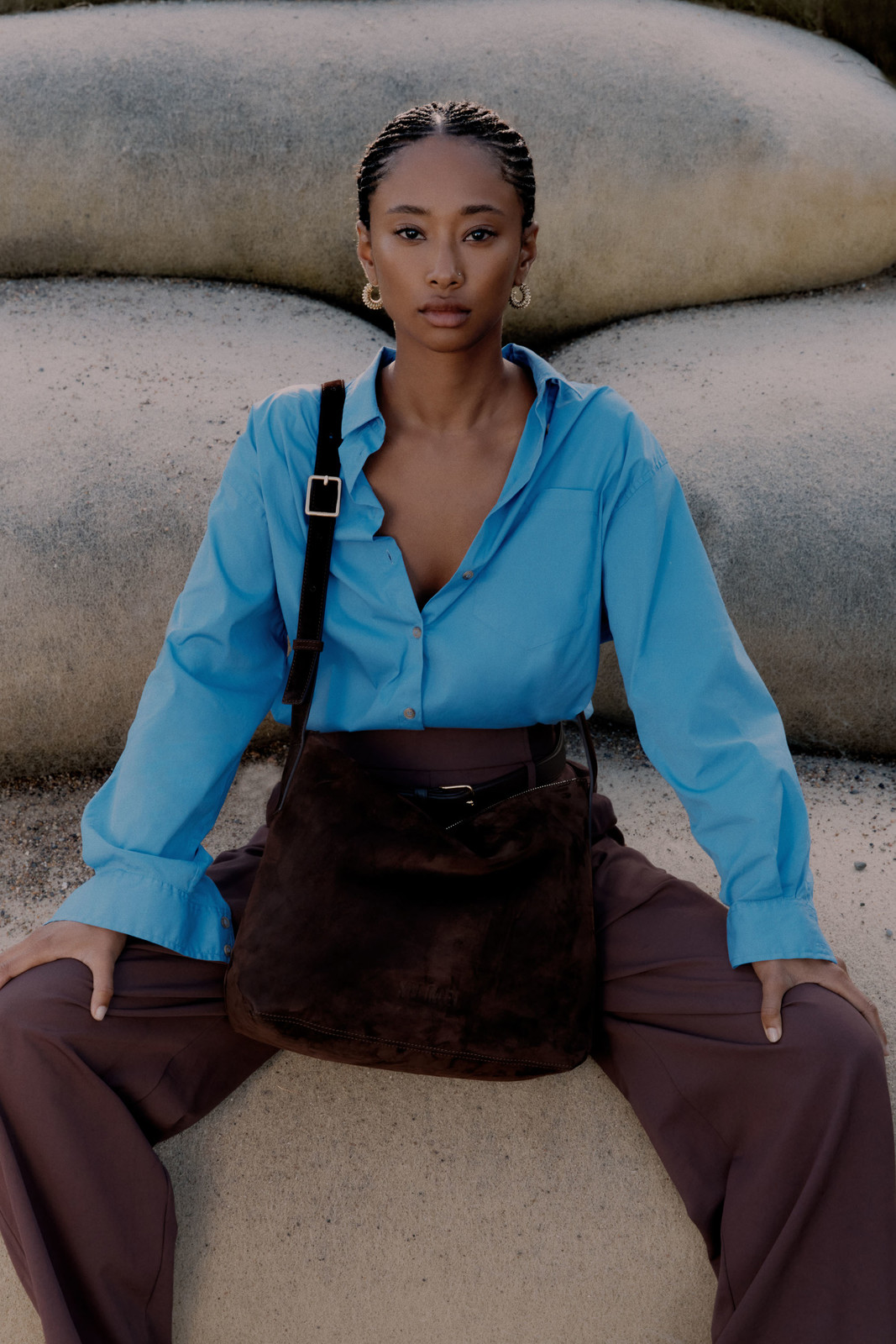 A woman with braided hair sits on a tan rock, wearing a blue shirt and brown pants, with a brown purse over her shoulder.