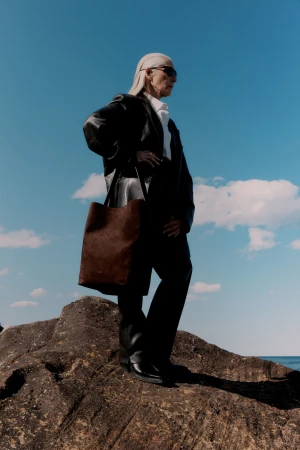 Elderly woman in black leather jacket and sunglasses holding a brown tote bag standing on a rocky outcrop under a clear blue sky