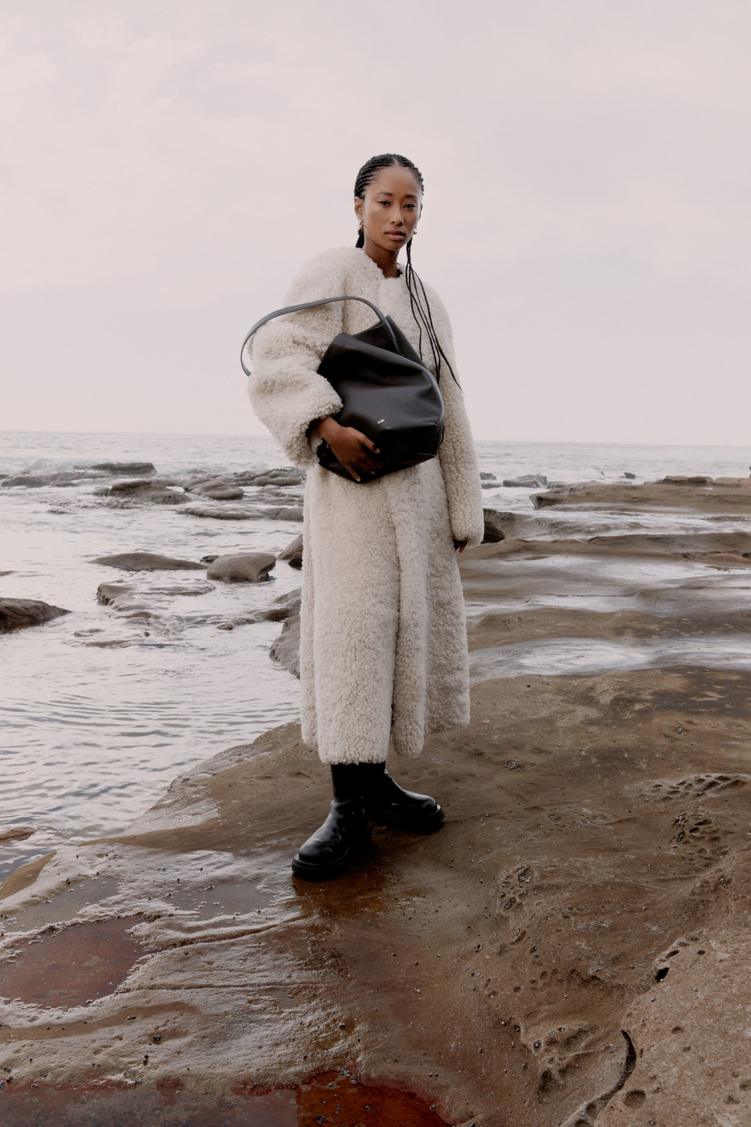 Fashion shot of a Black woman in a cream coat holding a black bag, standing on a rocky shore near the ocean under an overcast sky.