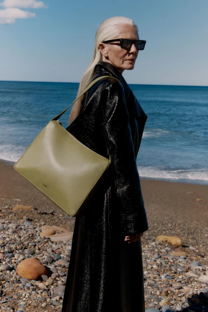 Elegant silver-haired woman wearing black coat poses with green tote bag on pebble beach with blue ocean and sky in lifestyle fashion shot.