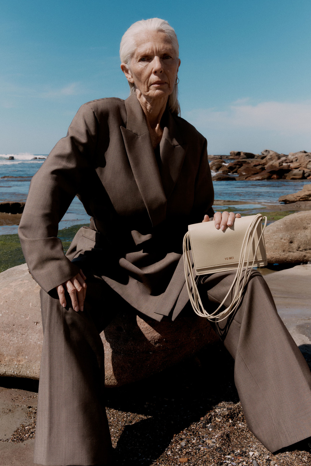 Older woman with silver hair wearing a loose brown suit holding a cream leather handbag on rocky coastal shore