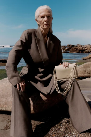 Older woman with silver hair wearing a loose brown suit holding a cream leather handbag on rocky coastal shore
