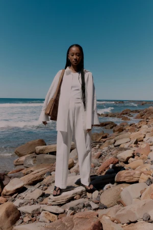 Woman in white shirt and trousers standing on rocky shore carrying a brown leather shoulder bag against ocean backdrop