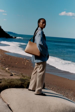 A tan purse is modeled by an African American woman in a blue jacket and tan slacks standing on a rock at a beach, sea, and headland.