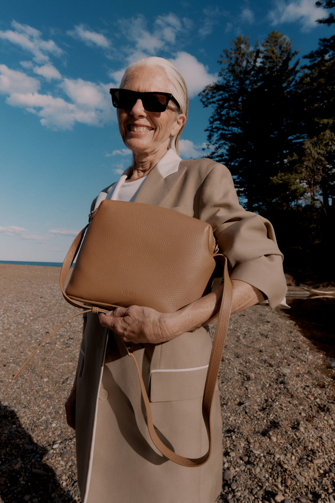 Elderly woman wearing sunglasses holding a tan leather crossbody bag on a beach