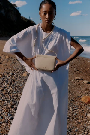 Model in a white dress holding a crossbody handbag on a sandy beach under a blue sky