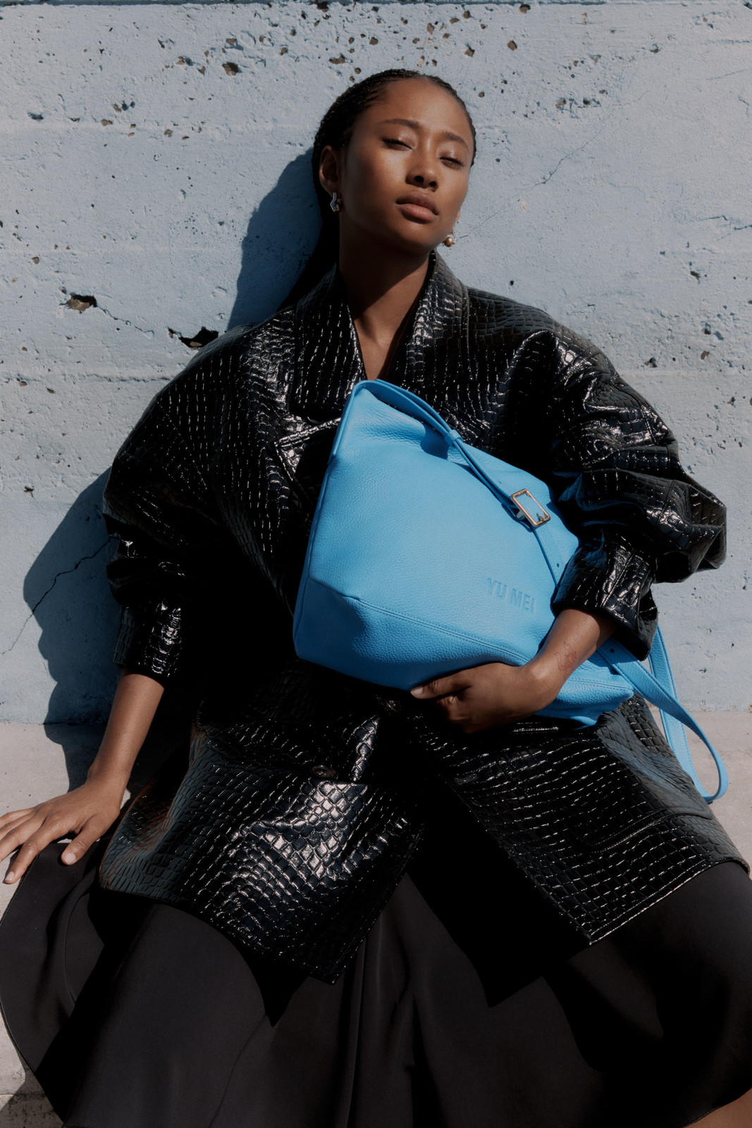 An adult woman poses with a blue handbag against a textured wall, wearing a black reptile-print jacket and black skirt in outdoor light.