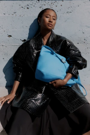 An adult woman poses with a blue handbag against a textured wall, wearing a black reptile-print jacket and black skirt in outdoor light.