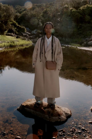 Model wearing a long beige trench coat standing on a large rock in the middle of a shallow river stream.