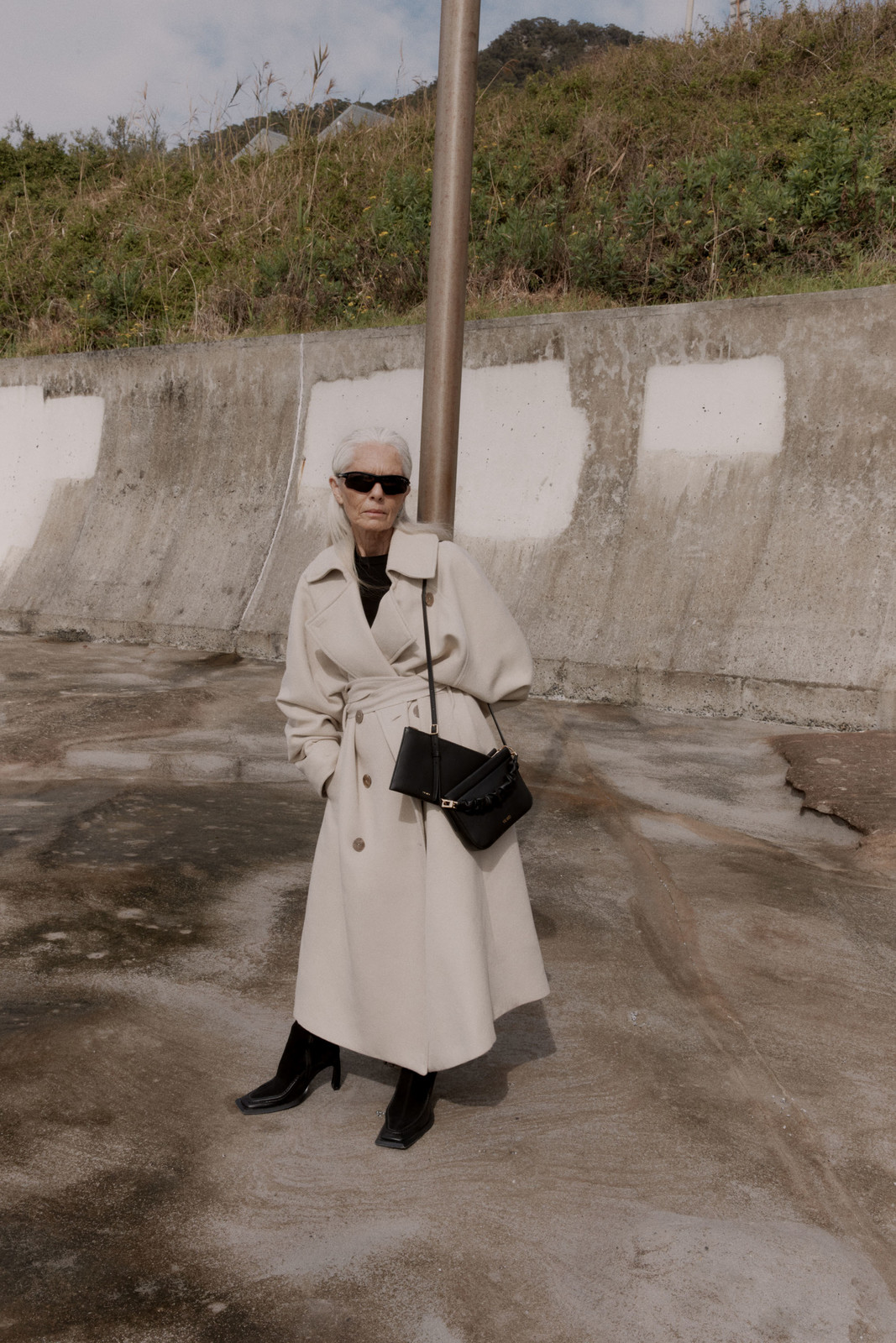 Elderly woman wearing a cream coat and black handbag standing outdoors near a concrete wall for Yu Mei SS 23 campaign