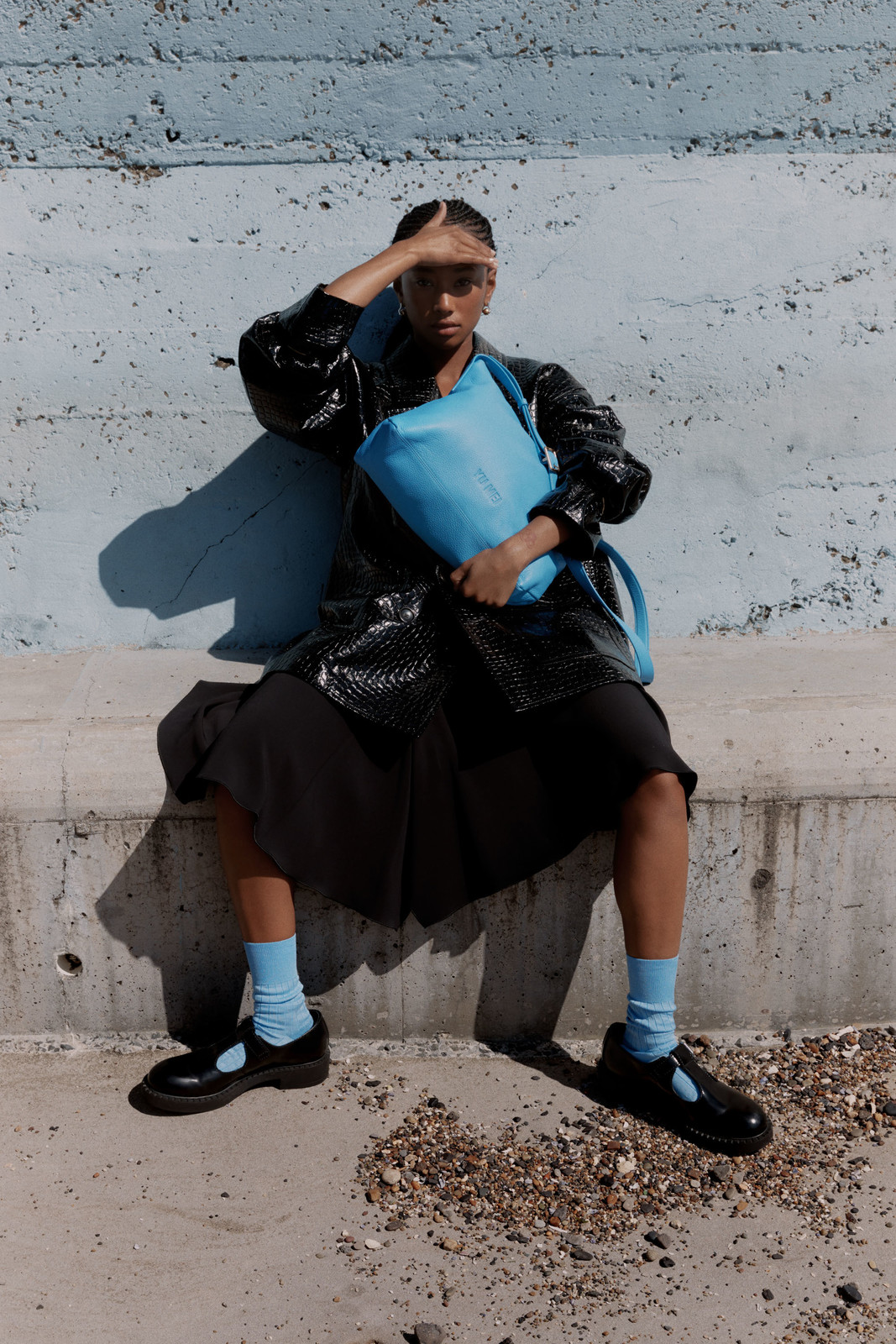 A person sits outdoors on a concrete ledge with a blue bag, wearing a black jacket, dress, and Mary Jane shoes with light blue socks.