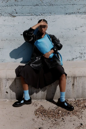 A person sits outdoors on a concrete ledge with a blue bag, wearing a black jacket, dress, and Mary Jane shoes with light blue socks.