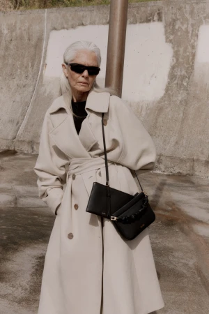 Older woman with long silver hair wearing dark sunglasses and an oversized cream coat, carrying black leather handbags against a textured wall