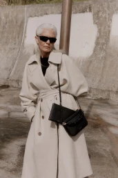 Elderly woman with silver hair wearing sunglasses and a cream trench coat carrying black handbags outdoors.