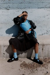 Model seated on a concrete ledge holding a bright blue leather handbag, wearing a glossy black jacket and blue socks.