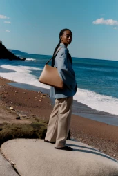 Model standing on a coastal rock holding a tan leather shoulder bag wearing an oversized blue blazer and beige trousers