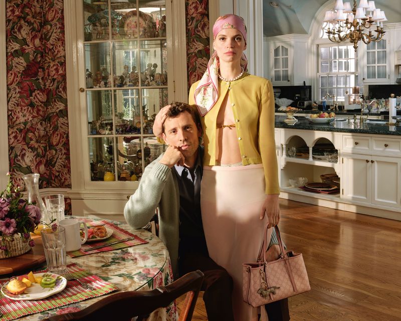 Indoor portrait of a man and woman; the man is seated at a table with food, and the woman is standing next to him with a handbag.